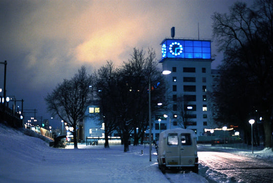 Picture of a street covered in snow with a old van in foreground. In backgroud Klokgebouw building.